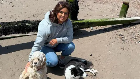 A woman crouching on the floor on a sandy beach, with two dogs sat near her. She is looking at the camera and smiling.