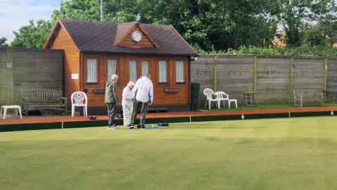 BBC/LIZ SAUL The green and club buildings of Halls Bowling Club, showing a red brick building and several white plastic chairs outside it. Three bowls players stand in front of the building, looking down at some bowls and a jack on the grass.