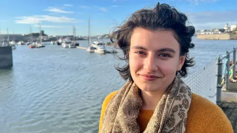 Anahita Laverack is standing next to the water at Plymouth harbour and is wearing a mustard jumper and a beige patterned scarf.