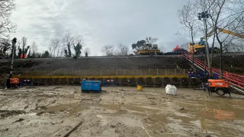 A view from the ground of where the landslip was and the embankment has been rebuilt, with machinery and people in hi-viz suits on train tracks.