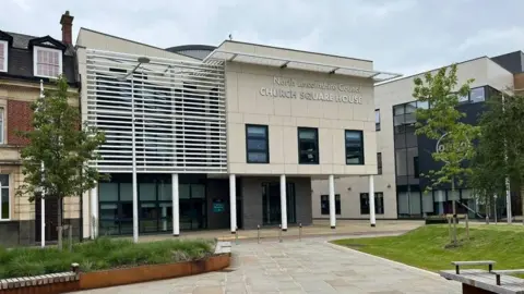 North Lincolnshire Council's building is a light, cream coloured building with various white poles and other buildings surrounding it.