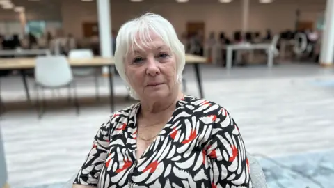 Kate Bradbrook/BBC Danielle Stone is wearing a black, white and red top and is sitting in a large indoor area with wooden tables and white chairs.