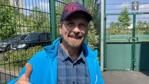 A man wearing a blue baseball cap with a red pattern and a blue check shirt under a blue fleece gives a thumbs-up to the camera.