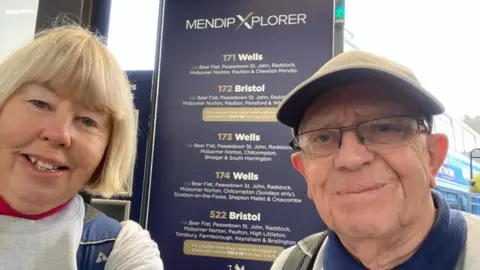 Kerstin Benham Mrs Banham, with blonde hair and wearing a light-coloured top with a blue backpack, and Mr Banham, wearing a light-coloured cap. They are standing in front of a notice showing bus routes in Somerset.