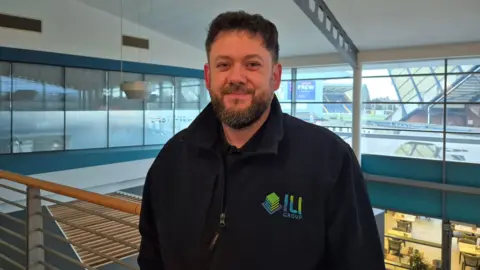 Greig Templeton is standing against an upstairs guardrail. He has a beard and moustache, short, dark hair and a black jacket with ILI Group written on in green and blue lettering.