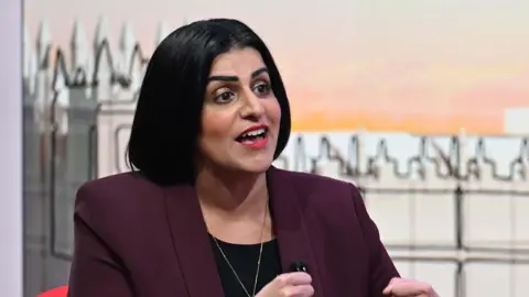 Home Secretary Shabana Mahmood wearing a maroon jacket over a black top. Her mouth is open as she talks. Behind her is a TV set with an illustration of the Houses of Parliament on it.