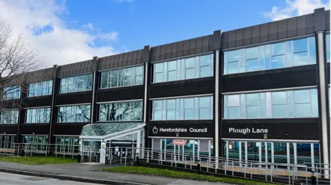 The exterior of a large three-storey building, with the signs "Herefordshire Council" and "Plough Lane" on the side of it. It has long, rectangular glass windows with dark cladding in between.