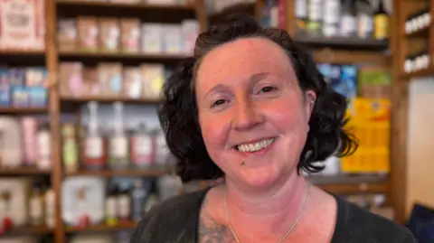 Johann smiles for the camera. She has short black hair and wears a black top. In the back ground, wooden shileves are neatly stacked with bottles and packets of different produce.