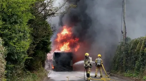 Two firefighters in brown clothing and yellow helmets are holding yellow hoses. A burning object on a road is behind them on the photo and there are bushes either side of the road.