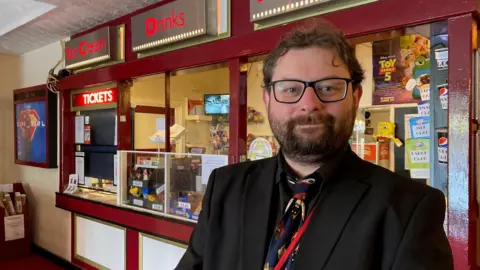 Charles Heslett/BBC A man with glasses and a dark beard and wearing a black suit and colourful tie stands in front of a old-style cinema ticket office painted in maroon and cream 