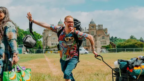 A man poses for the camera with his tongue out and an arm stretched in the air above him. He is pulling wheeled cart full of camping gear. It is a sunny picture with the imposing golden-stone Charlton Park country house behind.