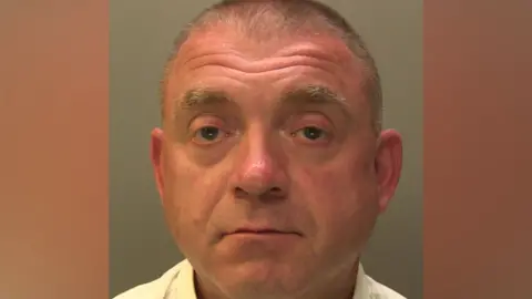Police mugshot of a man with short hair. He is wearing a yellow shirt and is looking directly at the camera.