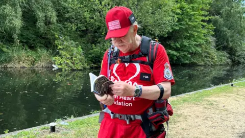 Supplied David stands in front of a canal. He is wearing a rucksack and reading a book. The title of the book is "The Grand Union Canal Walk"