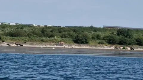 A lake can be seen looking out at a grassy bank across the other side with rocks below where people are congregated.