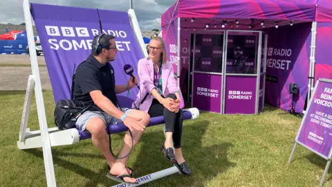 A man interviews a woman wearing a pink blazer on an oversized deck chair with the words "BBC Radio Somerset" written on it. In the background a purple and pink marquee, a sign board and a makeshift radio studio can be seen with the same words written on it.