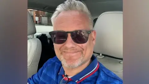 Supplied A selfie style image of Andy Cumbers. He is wearing sunglasses and a collared blue top. He is sitting in a car and smiling.