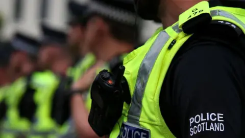 Six police officers standing in a line. They are all wearing black tops and florescent yellow vests. The faces of the officers in the background, who are all wearing hats, are blurred. The head of the officer who is photographed in foreground is cut off in the image. 