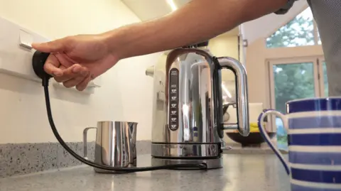 Getty Images A hand reaches out to switch on a kettle in a modern kitchen. There is a mug and a jug beside the kettle. 
