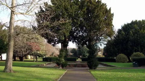Ewan Munro/Wikimedia Commons A scenic view of a paved pathway in Manor House Grounds, leading toward a cluster of large, dense evergreen trees. The path is flanked by well-maintained green lawns, with various deciduous trees and manicured hedges visible in the background. A small stone-bordered area sits at the end of the path, and a dark waste bin is positioned to the right of the walkway.