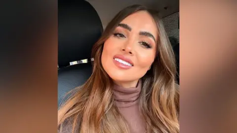 Jade Morgan has long brown hair and is wearing a brown polo neck jumper. She is sat in a car and is smiling at the camera.
