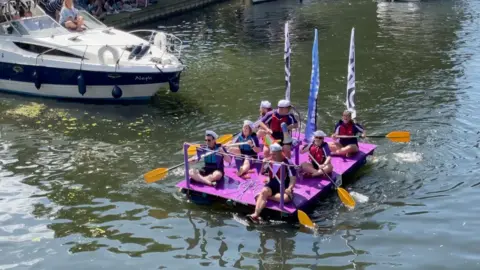 Seven people on a purple raft with flags on being rowed along the river