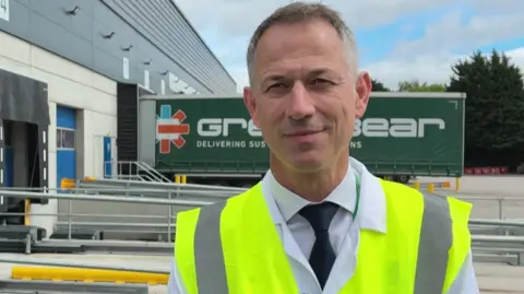 Mark Woodhead has short greying hair and is wearing a white shirt, black tie and high-vis yellow jacket. He is standing at the Port Sunlight distribution site with a lorry in the background.