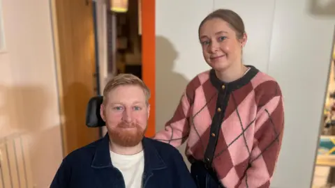 A man and woman pictured side by side. He is seated on the right, smiling at the camera, with ginger hair and beard. He is wearing a white t shirt and a dark open jacket. To his left is a young woman smiling at the camera, she has dark hair and is wearing a pink patterned cardigan.  