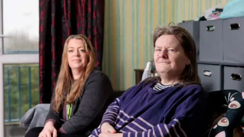 Image shows Jenny Devon, a support worker from Prima Housing who has long brown hair, sitting on the sofa next to Ruth. 
