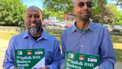 Two men stand smiling at the camera. they are outside and each holding a poster which reads 7th Suffolk BME Business Awards.