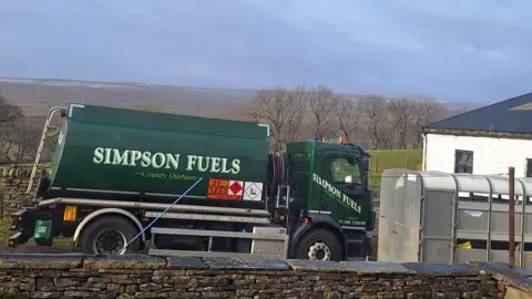 A Simpson Fuels tank is parked up outside the end of a white house which is partly obscured by a horsebox. 