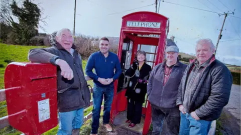 Mark McMillan Five residents in front of a red phone box. A man on the far left has his elbow placed on a red post box, he is wearing a wax jacket and light blue jeans. The man second from left is wearing a blue half zip top, dark blue jeans and light brown shoes. The woman in the middle is inside the phone box holding the phone to her ear, she is wearing a black coat and trousers. The man second from right is wearing a flat cap, purple fleece zip jacket and dark trousers. The man on the far right has white hair, wearing a black puffer jacket and blue jeans.