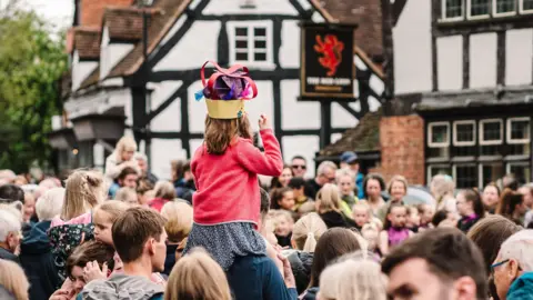 Visit Knowle A girl sits on a man's shoulders among a large crowd of people gathered in a village centre.