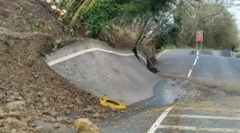 BBC One side of a road has been pushed up by a landslip under it and it resembles a skate ramp. In the distance, a large dip can be seen in another section of the route alongside a large crack in the road's surface.