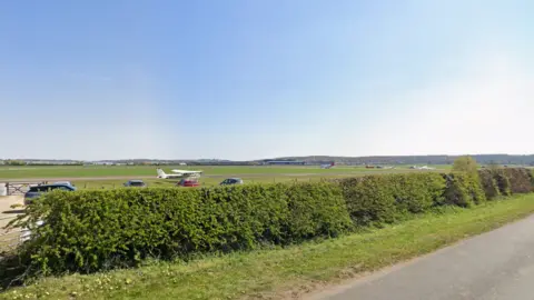 Google A relatively empty and large field with a few cars and a small white plane in it. In the foreground is a hedge and a strip of grass next to a road