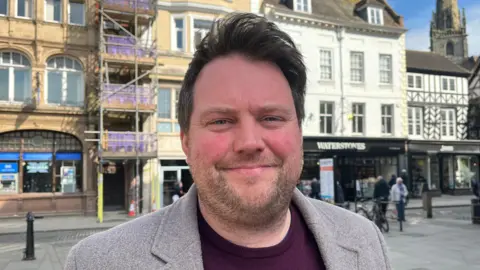 Seb is looking into the camera and smiling; he's wearing a maroon-coloured top with a grey wool blazer over the top. He's stood on a street in Shrewsbury, with a Waterstones and Halifax behind him, and a church spire in the distance. The sun is out and the sky is blue and clear. 