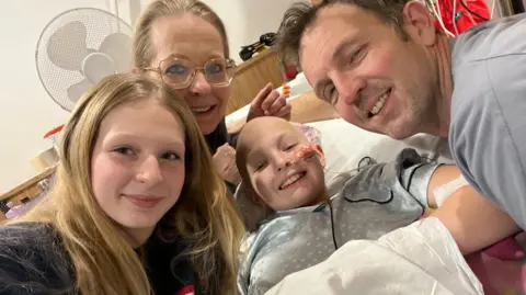 Millie is wearing a grey nighty while lying in bed. Her parents are leaning over her and her sister Jessica appears to be taking the photo.