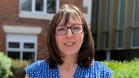 Head teacher Rebecca Mahony. She has shoulder-length dark hair and glasses and is wearing a blue blouse. She is standing in front of a school building.