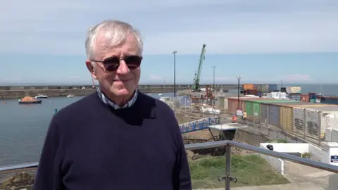 A man with sunglasses and grey hair smiling at the camera. He is wearing a blue jumper. In the background is a harbour.