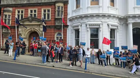 Crowds of protestors fill the pavement from left to right holding placards and flags. They are standing outside Tynwald, an ornate brick building behind on the left, and a circular white building on the right.