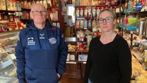 BBC/Seb Cheer A man wearing a Leeds Rhinos hoodie stands next to a woman wearing a black jumper. They are in a shop surrounded by old-style jars of traditional sweets.