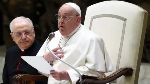 Reuters Pope Francis, sitting on a white chair, speaks into a microphone while holding papers. 