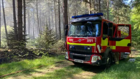 A fire engine parked in the middle of a forest. It is surrounded by trees. One of its doors is open as a firefighter leans inside.