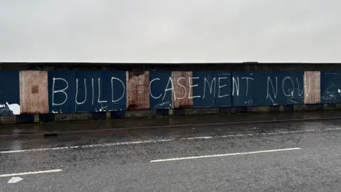 Boarded up wall that has been graffitied to say Build Casement Now, it runs alongside a road beside the stadium