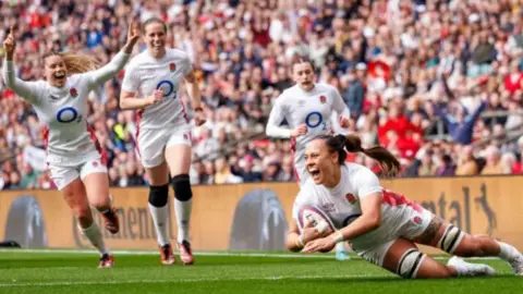 A woman in white England kit looks thrilled as she scores a rugby try with three other England players behind her looking equally happy