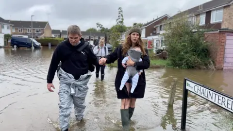 A family of three walks through flooded water in their street, Chaunterell Way, there are homes in the background and a black van to the left of the frame, the dad is standing to the left, he has short hair and wears a black hoodie with a Puma logo on the right of his chest and a silver waterproof boiler suit. He is holding a woman's hand, she wears a khaki green woollen beanie hat with a khaki green fur bobble on top of it and a black parka coat and green wellie boots, she carries a baby in a baby carrier.