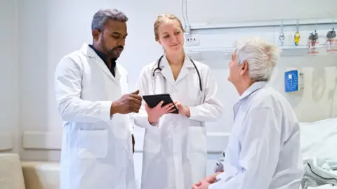 Two healthcare professionals wearing white coats, with one holding a tablet, talk to a patient with grey hair who is sitting on a hospital bed