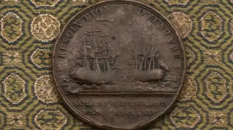 A medal with images of the ships used by Captain James Cook on his second voyage. The medal is sitting on a green and yellow patterned cloth.