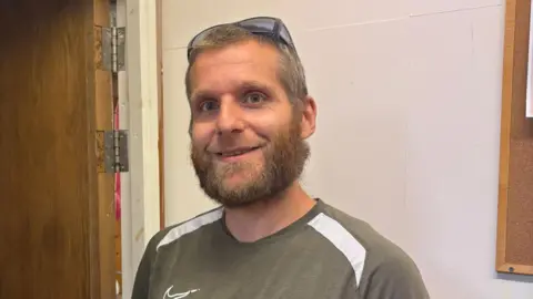 SFiCE A white man with brown hair and a beard. He is wearing a green striped t-shirt with sunglasses on his head.