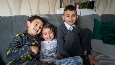 Tres jóvenes hijos sonrientes se sientan en un sofá en una sala de estar. Se agrupan de cerca. Una niña en el medio y un niño a su derecha lleva pijamas, mientras que un niño mayor a su izquierda lleva su uniforme escolar. Todos tienen cabello oscuro