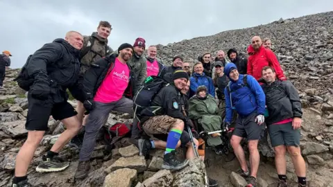 Pete A group of people stand on a mountain, including a boy in a wheelchair with Catherine, the Princess of Wales, stood next to him.